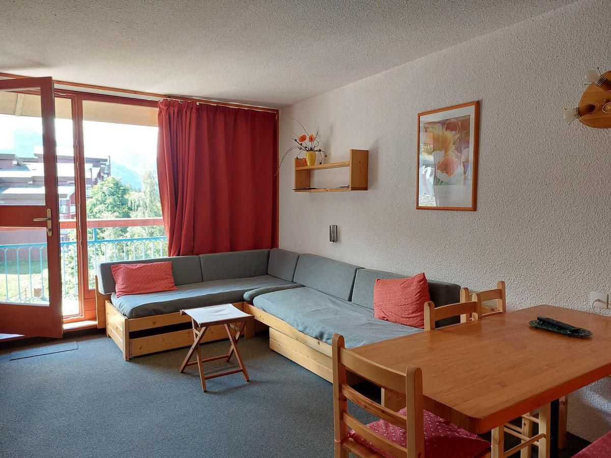 Een gezellige kamer met een bank, een klein tafeltje en een eettafel. Er is een raam met gordijn, een wandplank met een vaas, en een ingelijste foto.