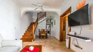A living room with a sofa, a coffee table with decorative items, a TV mounted on the wall, a staircase leading to another room, and a dining area with chairs and a table. - Casa Hannah - Marina Stroll by Villas Now (Playa Blanca)