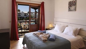 A hotel room with a bed, a nightstand, a lamp, a view of the pool and buildings outside. - Casa Hannah - Marina Stroll by Villas Now (Playa Blanca)