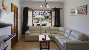 A living room with a sofa, a small table, a TV, and a window with a view of buildings and a patio. - Casa Hannah - Marina Stroll by Villas Now (Playa Blanca)