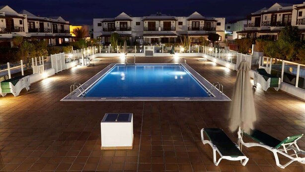 A swimming pool with lounge chairs and umbrellas, surrounded by buildings with balconies. - Casa Hannah - Marina Stroll by Villas Now (Playa Blanca)