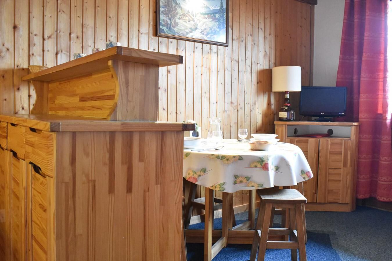 A wooden interior with a dining table set for two, a television on a wooden cabinet, and a painting on the wall.