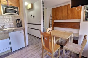 A compact kitchen with a microwave, coffee maker, and a small refrigerator. There is a wooden table with two chairs and a wooden cabinet above it.