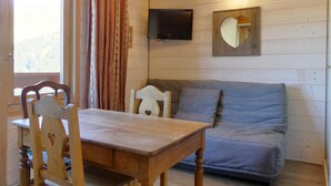 A cozy room with a wooden table and chairs, a grey sofa, a mounted TV, and a heart-shaped mirror on the wall. - 50m from ski slopes, 4th floor, 18m², Méribel-Mottaret (Méribel - Les Allues)