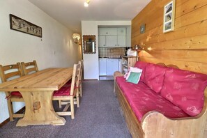A cozy room with a wooden dining table and chairs, a red sofa, a kitchen area, and a mirror on the wall.