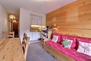 Ein gemütliches Zimmer mit Holzwänden, einem rosa Sofa mit gemusterten Kissen, einem Holzesstisch mit Stühlen und einer Küchenzeile mit weißen Schränken und einem Spülbecken.
