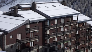 A modern multi-story building with a snow-covered roof, balconies, and a stone base. - 50m from ski slopes, Ground floor, 14m², Méribel-Mottaret (Méribel - Les Allues)