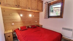 A bedroom with a wooden headboard, a red bedspread, and a window with a view of a building. - 50m from ski slopes, 1st floor, balcony, fireplace or stove, 45m², Méribel-Mottaret (Méribel - Les Allues)