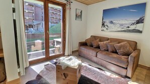 A living room with a brown leather sofa, a wooden chest, and a view of a mountainous landscape through a large sliding glass door. - 50m from ski slopes, 1st floor, balcony, fireplace or stove, 45m², Méribel-Mottaret (Méribel - Les Allues)