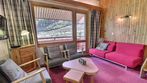 A cozy room with a red sofa, two armchairs, a small table, and a window offering a view of a forested area. - 10m from ski slopes, 3rd floor, balcony, fireplace or stove, 55m², Méribel-Mottaret (Méribel - Les Allues)