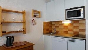 Een kleine keuken met witte kastjes, een magnetron, een spoelbak en een houten plank met glazen.