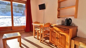 Een knus ingerichte kamer met een houten tafel, stoelen, een klein kastje, een televisie en uitzicht op een besneeerd landschap door een groot raam.
