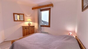 A bedroom with a bed, a wooden dresser, a mirror, a window with a view of a mountain, and a lamp. - 10m from ski slopes, 1st floor, balcony, 49m², Méribel-Mottaret (Méribel - Les Allues)