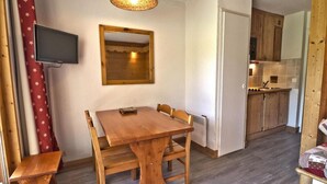 Um quarto aconchegante com uma mesa de jantar de madeira, cadeiras, uma TV montada na parede e uma área de cozinha com armários de madeira e painel de azulejos.