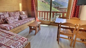 Quarto aconchegante com um sofá estampado, uma mesa de madeira e cadeiras, uma TV e uma grande porta de vidro deslizante com vista para as montanhas.