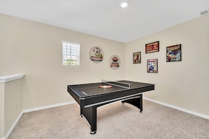 A room with a pool table, a window with blinds, and wall decorations including sports and superhero posters.