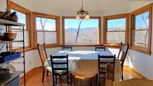 Ein Esszimmer mit großem Tisch, Stühlen und einer Kandelaber. Drei große Fenster mit Holzrahmen bieten Blick auf Bäume und Berge.