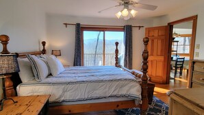 Ein Schlafzimmer mit einem hölzernen Bett, einem Nachttisch, einem Tisch, einem Deckenventilator und Blick ins Freie durch große Fenster.