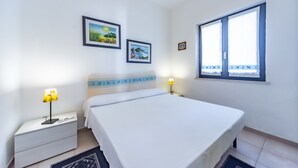 A bedroom with a bed, bedside tables, wall-mounted air conditioning, framed pictures, and a window with curtains.