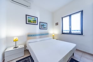 Quarto com cama, mesas de cabeceira, ar condicionado fixado na parede, quadros emoldurados e janela com cortinas.