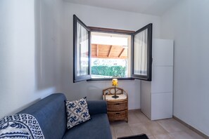 Um quarto pequeno com um sofá azul, uma mesinha de vime com uma luminária e uma janela com persianas abertas.