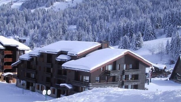 Un chalet à étages, dont le toit est recouvert de neige, entouré d’un paysage enneigé et d’une forêt dense.