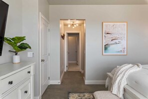 Un dormitorio con cama, mesita de noche, planta, cuadro enmarco y un pasillo que conduce a otra habitación.