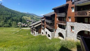 A row of chalet-style buildings with stone foundations and wooden balconies, set against a backdrop of green hills and a clear blue sky.