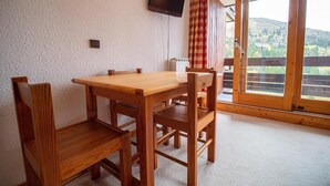 A wooden dining table with four chairs, a television mounted on the wall, and a large window with a view of hills.