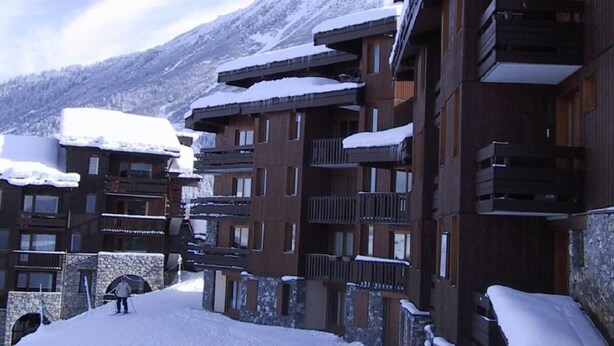 A snow-covered ski resort with multi-story buildings, ski slopes, and a mountainous backdrop.