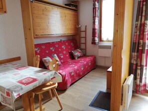 Une petite pièce chaleureuse, avec un lit superposé en bois, une table à manger recouverte d’une nappe à motifs et un canapé rouge orné de motifs en forme de cœur.