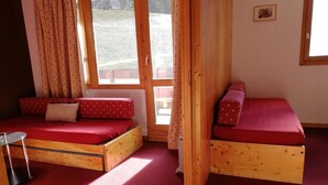 A room with two wooden beds, red bedding, a small table, and a window with curtains.