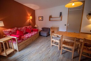 A cozy room with a wooden sofa, a dining table, and chairs. There is a small table and a chair near the sofa.