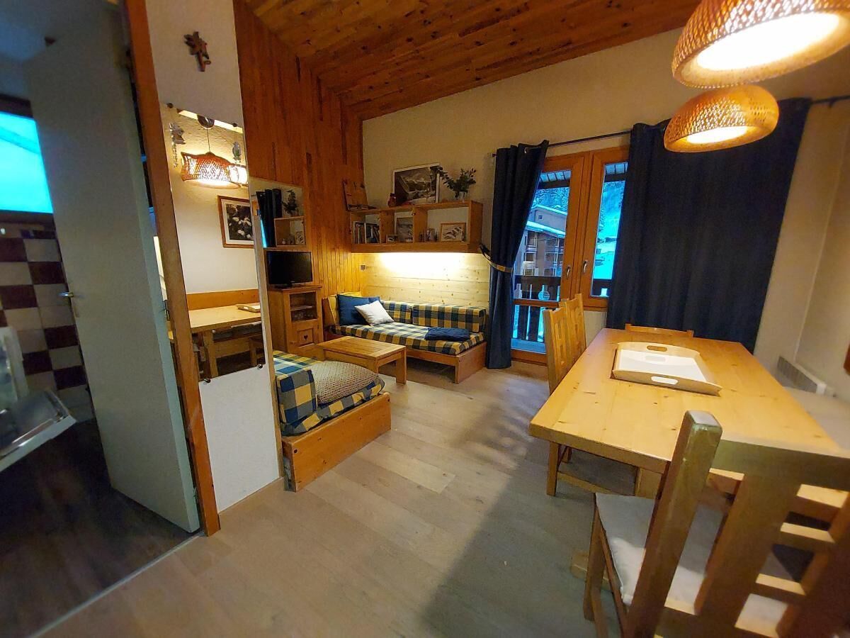 Ein gemütliches Zimmer mit Holzbalkendecke, einem Esstisch mit Stühlen, einer Couch und einer kleinen Küchenzeile mit Kühlschrank und Regalen.