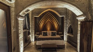 A bedroom with a stone archway, a bed with pillows, a bench, and a nightstand.