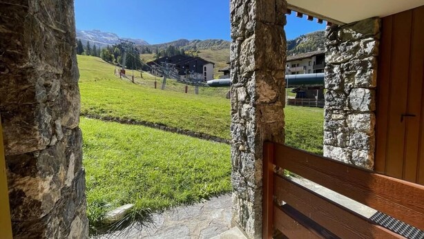 Un portico con cornice in pietra e una panca di legno, affacciato su una zona erbosa con edifici e montagne sullo sfondo.