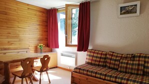 A cozy room with a wooden dining table, two wooden chairs, a plaid sofa, and a window with red curtains. - 100m from shops, 2nd floor, view mountain, balcony, tv, ski locker, 32m², Valmorel (Valmorel)