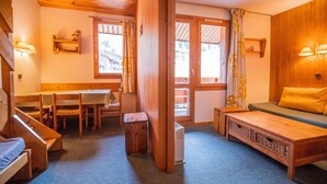 Et hyggeligt rum med en træbænk, en sofa, et bord og en stol. Der er vinduer med gardiner, en radiator og en vægmonteret hylde.
