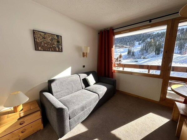 Ein gemütliches Zimmer mit grauem Sofa, einem hölzernen Nachttisch, einer Lampe und einem Fenster mit Blick auf eine verschneite Landschaft.