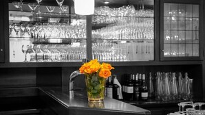 A bar area with a glass pitcher of orange roses, shelves of wine bottles, and various glasses. - Hotel-Restaurant Zum Schwanen (Wermelskirchen)