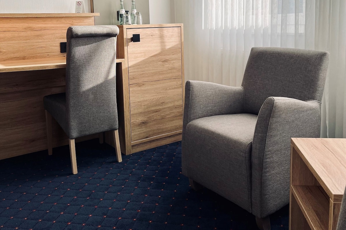 Habitació d'hotel amb escriptori de fusta, una butaca gris, una taula de fusta petita i una finestra gran amb cortines primes.