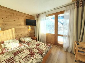 Quarto com parede de madeira, cama com roupa de cama estampada, TV fixada acima da cama e janela com cortinas.