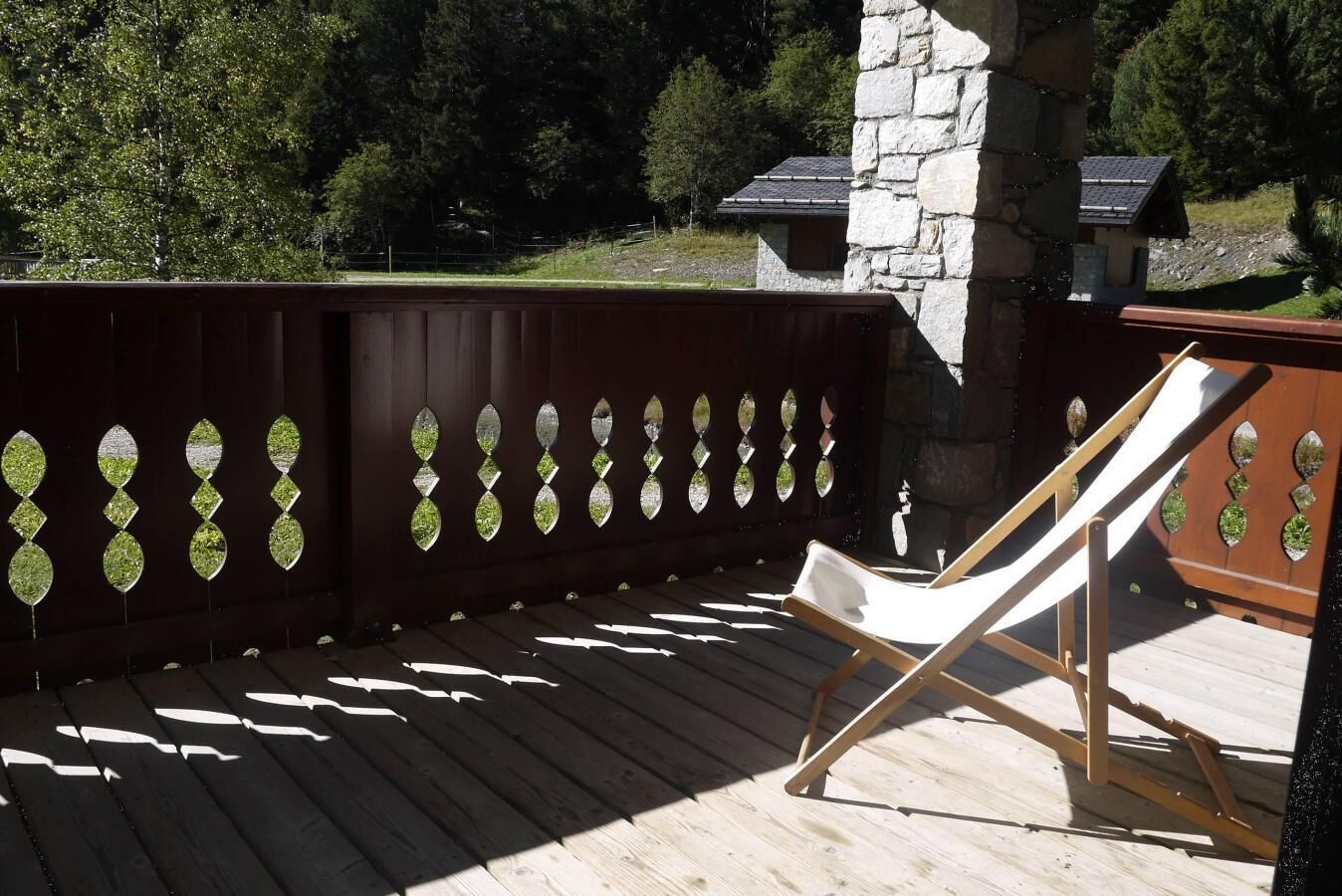 A wooden deck with a white lounge chair, a stone pillar, and a wooden fence with decorative glass panels.