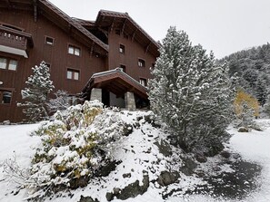 A multi-story wooden building with a stone entrance, surrounded by snow-covered trees and shrubs.