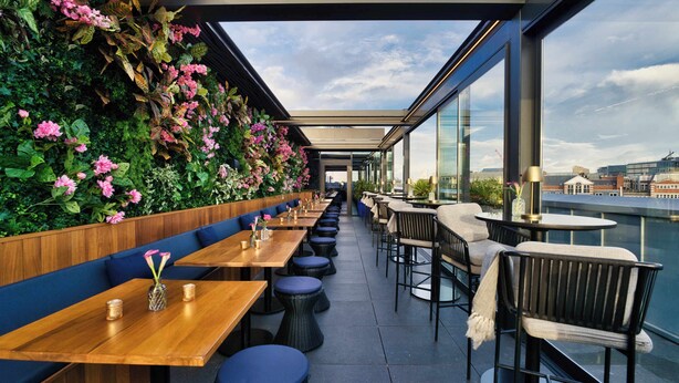An outdoor dining area with wooden tables and blue cushioned seating, a green wall with flowers, and a city view. - AMANO Covent Garden (London)