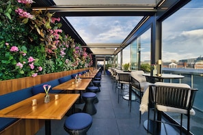 An outdoor dining area with wooden tables and blue cushioned seating, a green wall with flowers, and a city view. - AMANO Covent Garden (London)