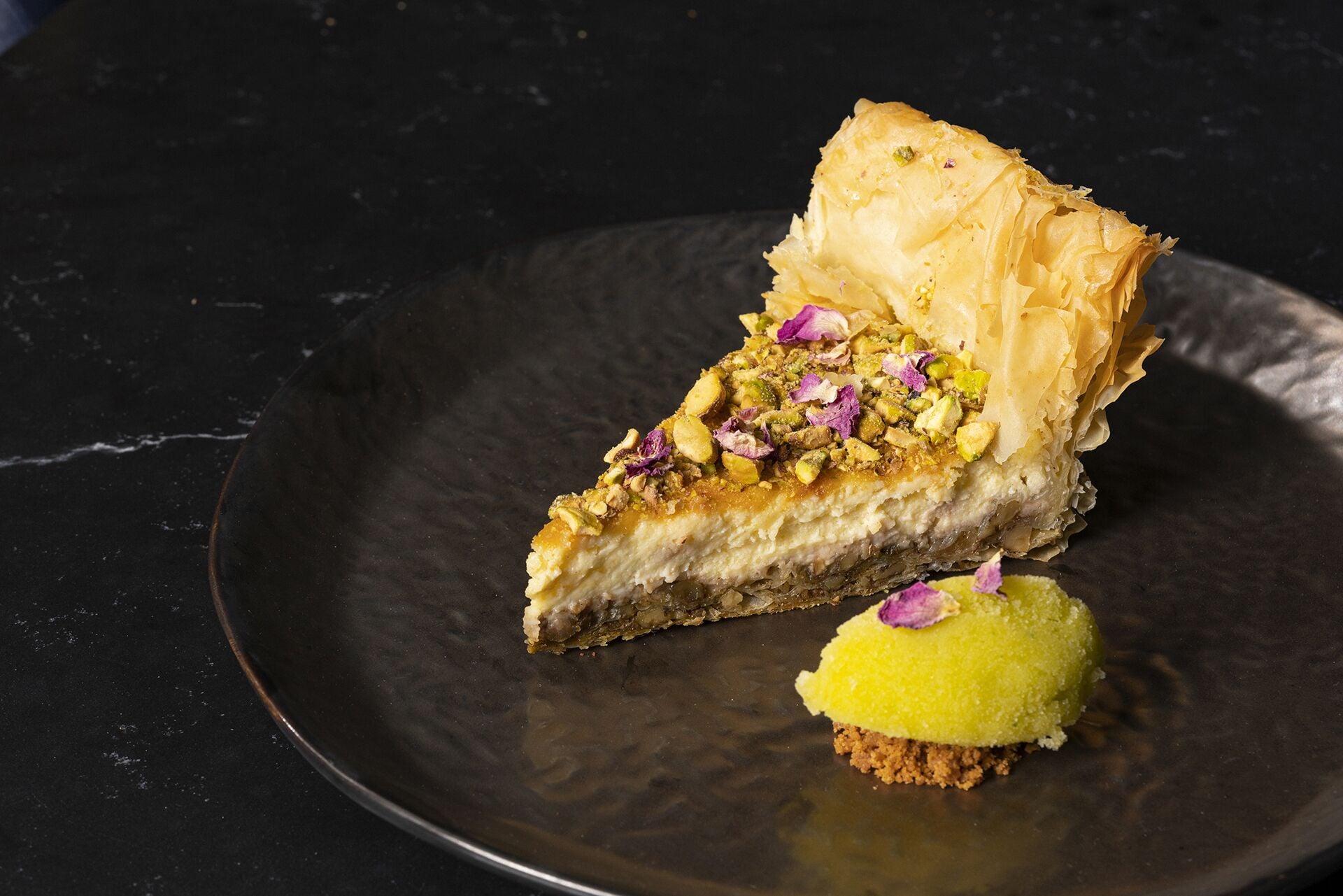 A slice of layered pastry with nuts and a small round pastry on a dark plate.