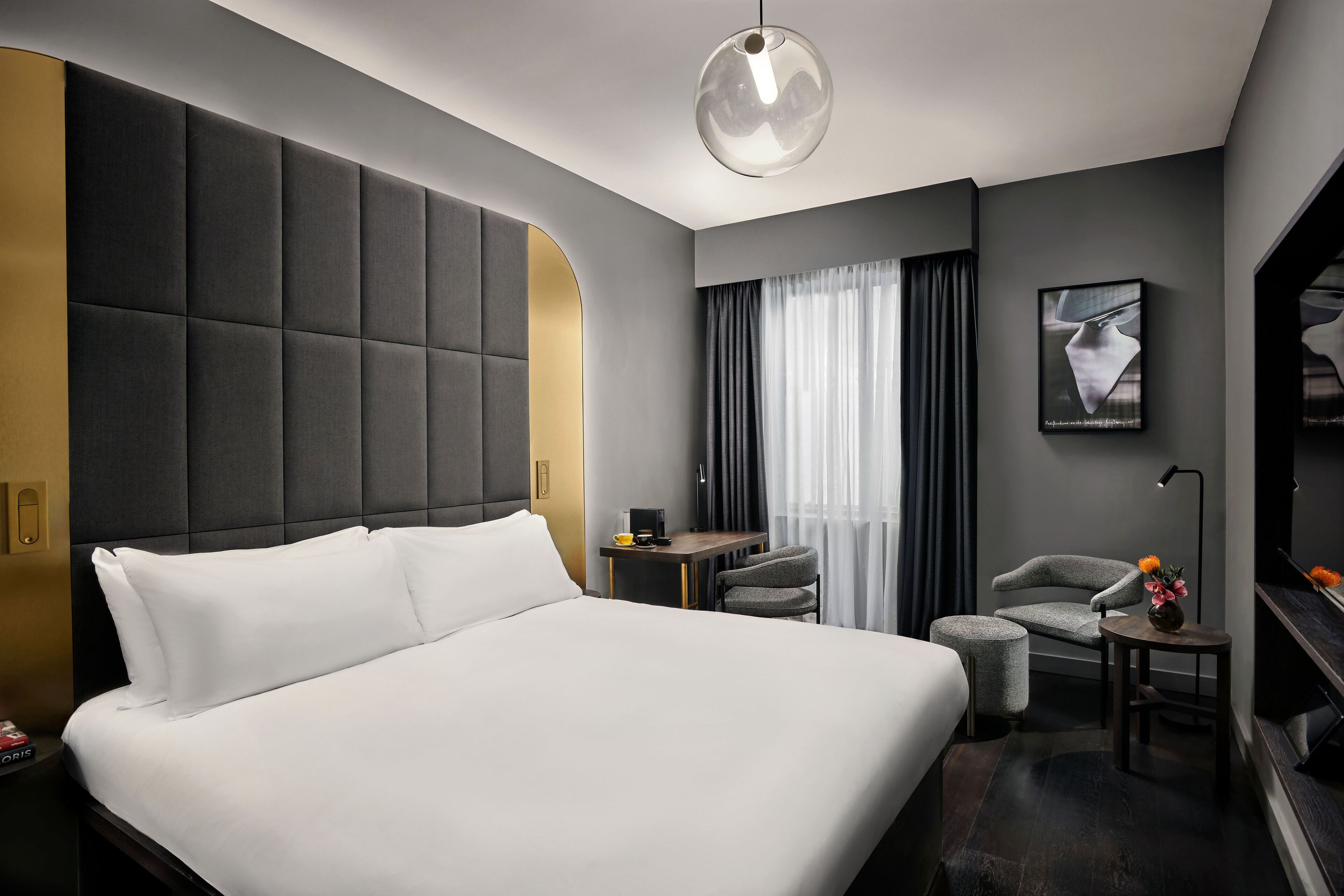Quarto de hotel moderno com uma cama grande, uma escrivaninha, uma cadeira e uma pequena mesa com flores.
