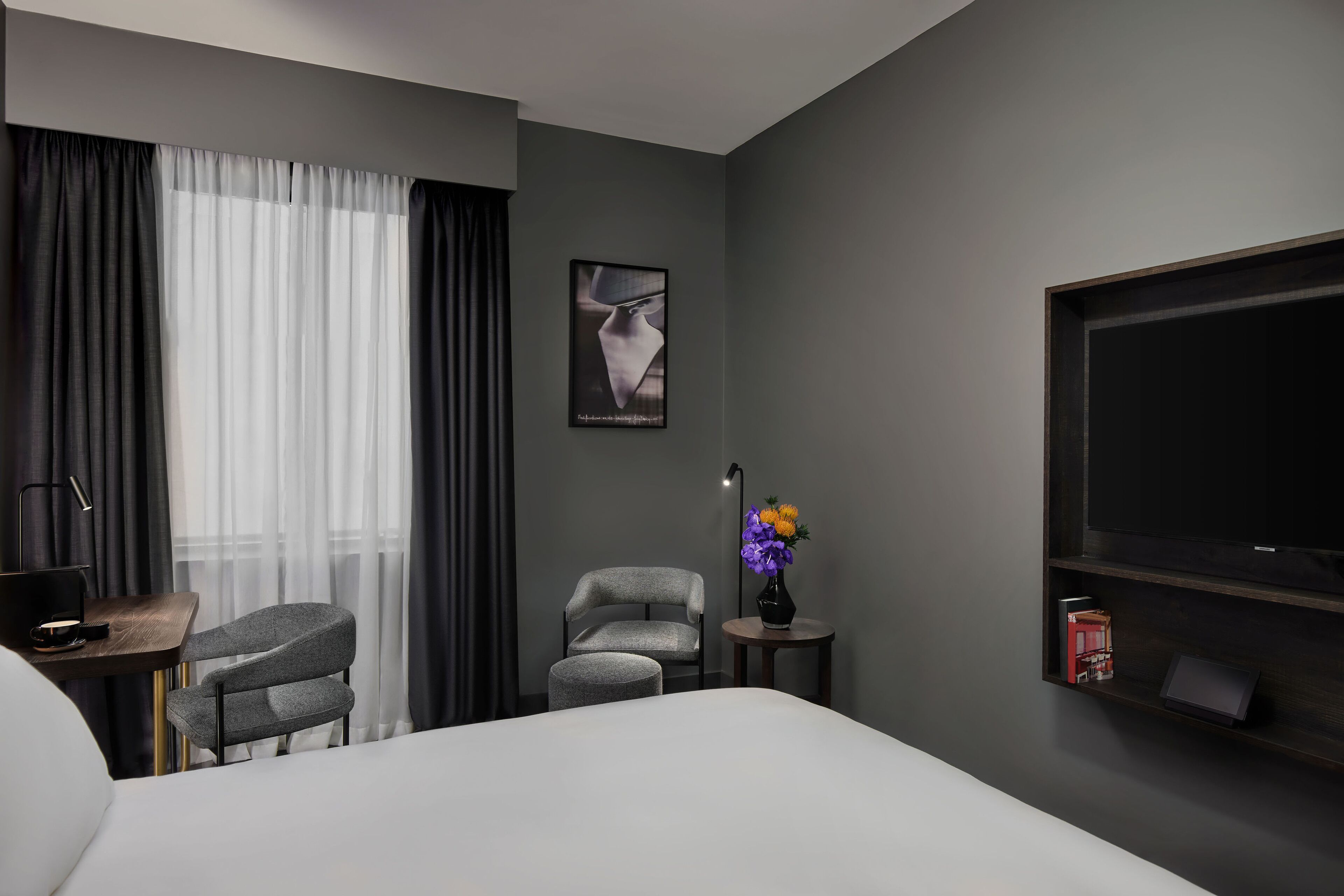 Une chambre d’hôtel moderne avec un grand lit, deux fauteuils gris, une petite table avec une lampe, un téléviseur posé sur une étagère en bois et un tableau encadré au mur.