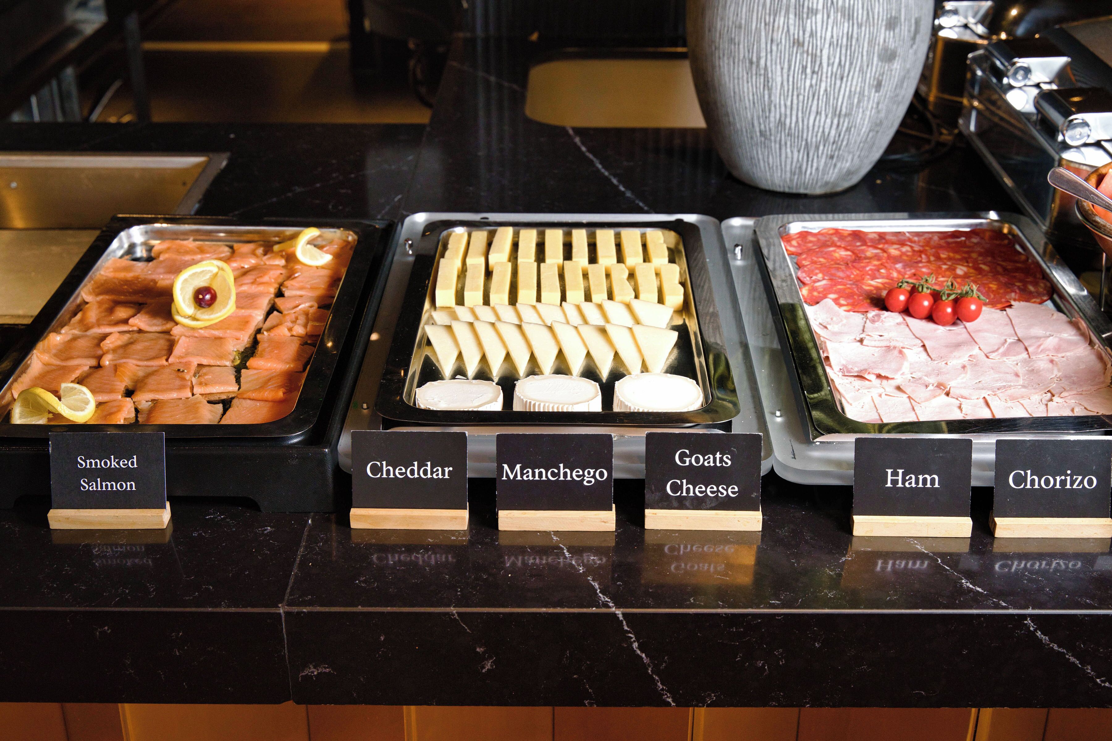 A buffet with labeled trays of smoked salmon, cheddar, manchego, goats cheese, ham, and chorizo.